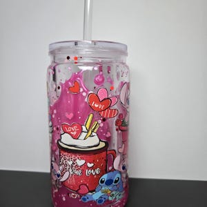 K&ouml;nnte beinhalten: Transparenter Kunststoffbecher mit Deckel und Strohhalm. Der Becher hat ein rosa-rotes Design mit Herzen, dem Wort "Love" und Zeichentrickfiguren. Der Becher ist mit rosa Glitzer und Fl&uuml;ssigkeit gef&uuml;llt.