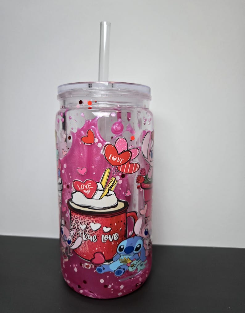 K&ouml;nnte beinhalten: Transparenter Kunststoffbecher mit Deckel und Strohhalm. Der Becher hat ein rosa-rotes Design mit Herzen, dem Wort "Love" und Zeichentrickfiguren. Der Becher ist mit rosa Glitzer und Fl&uuml;ssigkeit gef&uuml;llt.