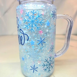 May include: A clear plastic tumbler with a handle and lid, filled with silver glitter and blue liquid. The exterior features blue snowflake and star decals, along with partial blue cursive text on the side of the cup.