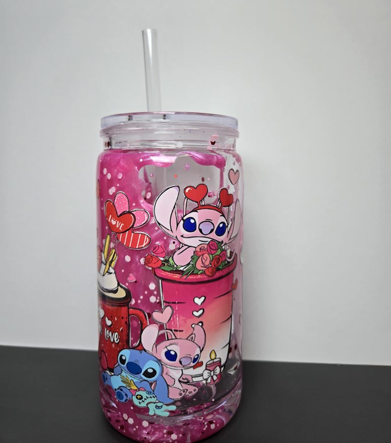 K&ouml;nnte beinhalten: Ein klarer Becher mit einem rosa-wei&szlig;en Design mit Zeichentrickfiguren, Herzen und dem Wort "Love". Der Becher enth&auml;lt einen klaren Strohhalm und einen klaren Deckel. Der Becher ist mit rosa Glitzer gef&uuml;llt.