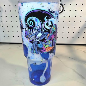 May include: A clear plastic tumbler featuring a graphic of Jack Skellington and Sally from The Nightmare Before Christmas. The design includes a white and blue drip effect with iridescent glitter accents throughout the body of the cup.