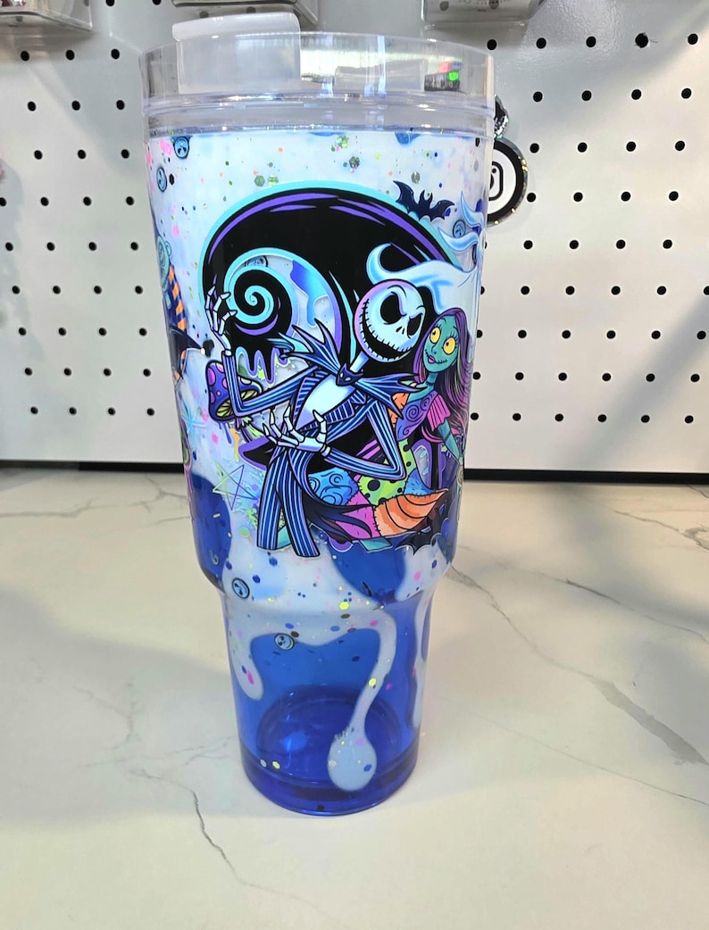 May include: A clear plastic tumbler featuring a graphic of Jack Skellington and Sally from The Nightmare Before Christmas. The design includes a white and blue drip effect with iridescent glitter accents throughout the body of the cup.