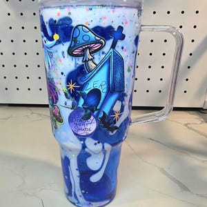 May include: A clear plastic tumbler with a handle and lid, featuring a blue and white swirling liquid effect with colorful glitter. The exterior displays graphics of a blue dog house, a mushroom, a bat, and a bottle labeled Deadly Night Shade.
