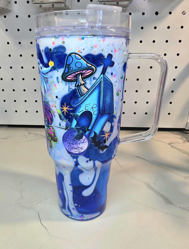 May include: A clear plastic tumbler with a handle and lid, featuring a blue and white swirling liquid effect with colorful glitter. The exterior displays graphics of a blue dog house, a mushroom, a bat, and a bottle labeled Deadly Night Shade.