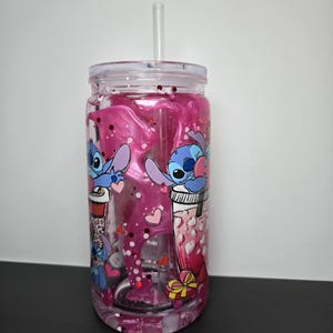 K&ouml;nnte beinhalten: Ein klarer Kunststoffbecher mit Deckel und Strohhalm, gef&uuml;llt mit rosa Fl&uuml;ssigkeit und Glitzer. Der Becher ist mit Cartoon-Bildern von Stitch aus Lilo & Stitch, Herzen und Geschenkboxen verziert. F&uuml;r kalte Getr&auml;nke.