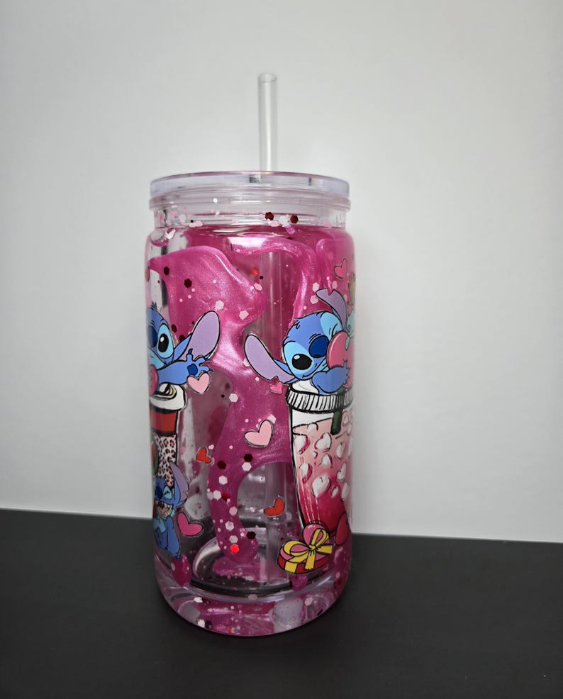K&ouml;nnte beinhalten: Ein klarer Kunststoffbecher mit Deckel und Strohhalm, gef&uuml;llt mit rosa Fl&uuml;ssigkeit und Glitzer. Der Becher ist mit Cartoon-Bildern von Stitch aus Lilo & Stitch, Herzen und Geschenkboxen verziert. F&uuml;r kalte Getr&auml;nke.