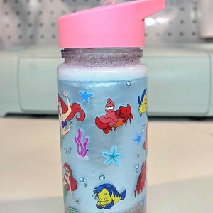 Może przedstawiać: Przezroczysta butelka na wodę z r&oacute;żową pokrywą z klapką. Butelka ma jasnoniebieskie tło z kresk&oacute;wkowymi obrazkami Arielki, Florka, Sebastiana i innych stworzeń morskich. Dno butelki ma warstwę szarego piasku.