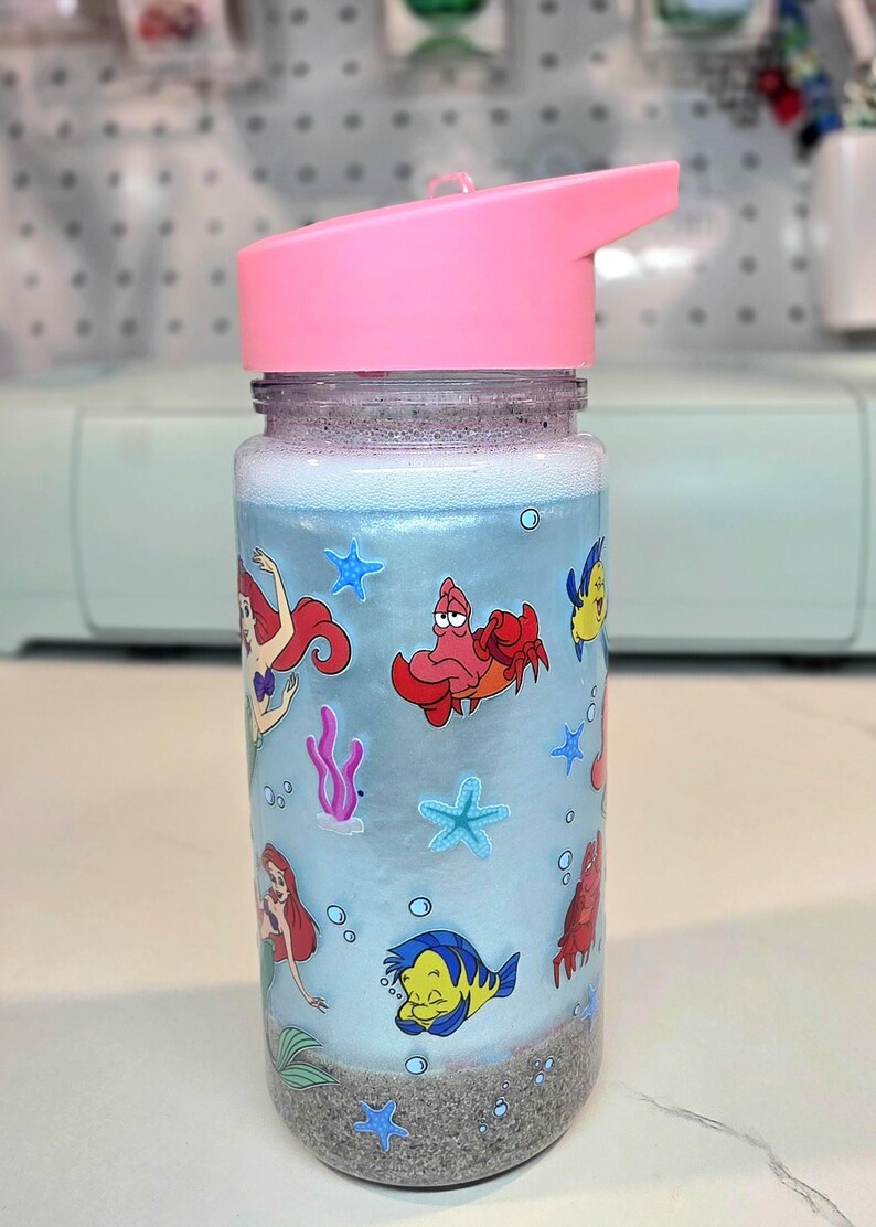 Może przedstawiać: Przezroczysta butelka na wodę z r&oacute;żową pokrywą z klapką. Butelka ma jasnoniebieskie tło z kresk&oacute;wkowymi obrazkami Arielki, Florka, Sebastiana i innych stworzeń morskich. Dno butelki ma warstwę szarego piasku.