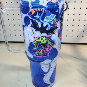 May include: A clear blue tumbler with a handle and a lid, featuring a decorative wrap with white swirling patterns, colorful glitter, and graphic illustrations of a green mushroom, a black bat, and a blue skull character with orange stripes.