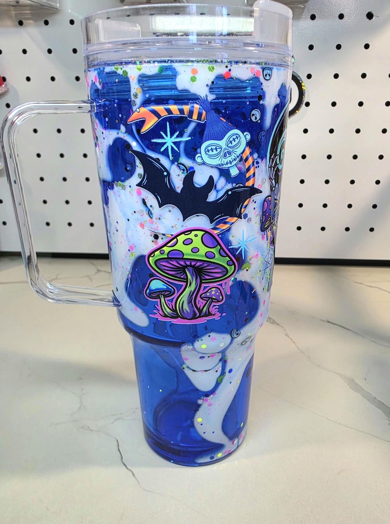 May include: A clear blue tumbler with a handle and a lid, featuring a decorative wrap with white swirling patterns, colorful glitter, and graphic illustrations of a green mushroom, a black bat, and a blue skull character with orange stripes.