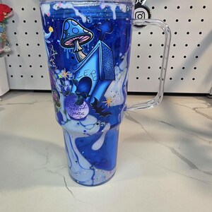 May include: A clear, double-walled tumbler with a handle and lid, featuring a blue liquid-filled design with white drip effects. The exterior displays a blue doghouse labeled Zero, a spotted mushroom, a bat, and a bottle labeled Deadly Night Shade.