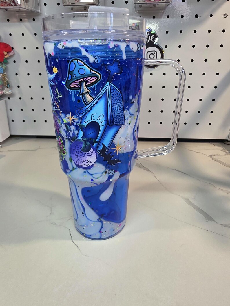 May include: A clear, double-walled tumbler with a handle and lid, featuring a blue liquid-filled design with white drip effects. The exterior displays a blue doghouse labeled Zero, a spotted mushroom, a bat, and a bottle labeled Deadly Night Shade.