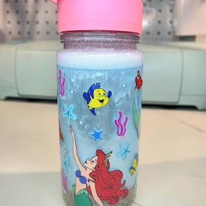 Może przedstawiać: Przezroczysta butelka na wodę z r&oacute;żową pokrywką, z motywem syreny i stworzeń morskich. Butelka ma jasnoniebieskie tło z ilustracjami Arielki, Florka i innych stworzeń morskich. Dno butelki ma wygląd piasku.