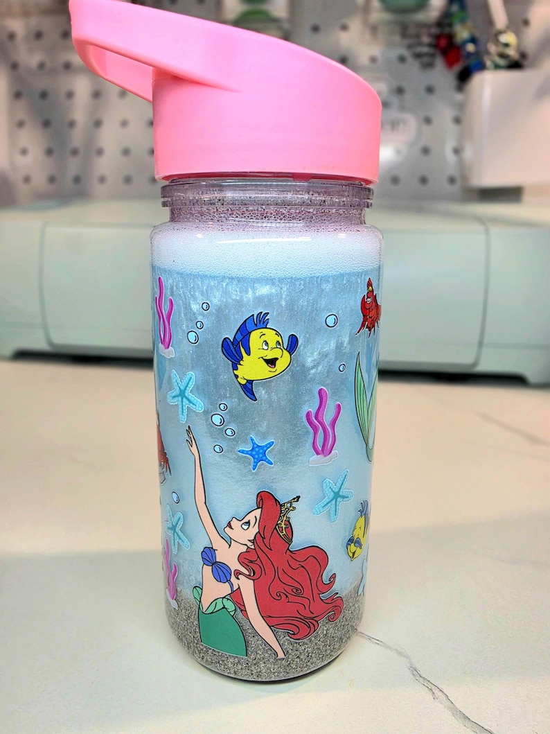 Może przedstawiać: Przezroczysta butelka na wodę z r&oacute;żową pokrywką, z motywem syreny i stworzeń morskich. Butelka ma jasnoniebieskie tło z ilustracjami Arielki, Florka i innych stworzeń morskich. Dno butelki ma wygląd piasku.