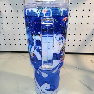 May include: A clear plastic tumbler with a handle, featuring a blue and white swirling liquid design with glitter. The exterior is decorated with colorful graphic illustrations of mushrooms, stars, and abstract shapes, topped with a clear lid.