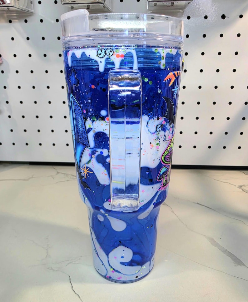May include: A clear plastic tumbler with a handle, featuring a blue and white swirling liquid design with glitter. The exterior is decorated with colorful graphic illustrations of mushrooms, stars, and abstract shapes, topped with a clear lid.