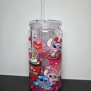 K&ouml;nnte beinhalten: Transparenter Kunststoffbecher mit Deckel und Strohhalm. Der Becher hat ein Design mit Zeichentrickfiguren, Herzen und dem Wort "love". Der Boden des Bechers ist mit rosa Glitzer gef&uuml;llt.