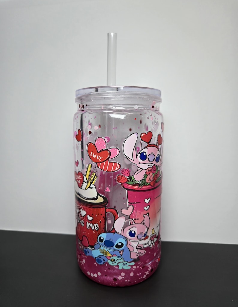 K&ouml;nnte beinhalten: Transparenter Kunststoffbecher mit Deckel und Strohhalm. Der Becher hat ein Design mit Zeichentrickfiguren, Herzen und dem Wort "love". Der Boden des Bechers ist mit rosa Glitzer gef&uuml;llt.
