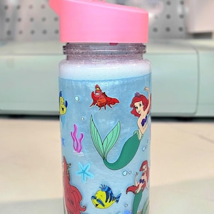 Może przedstawiać: Przezroczysta butelka na wodę z r&oacute;żową pokrywką z motywem syrenki. Butelka ma jasnoniebieskie tło z kresk&oacute;wkowymi ilustracjami syren, ryb i innych stworzeń morskich. Butelka jest wypełniona niebieskim płynem i piaszczystym dnem.