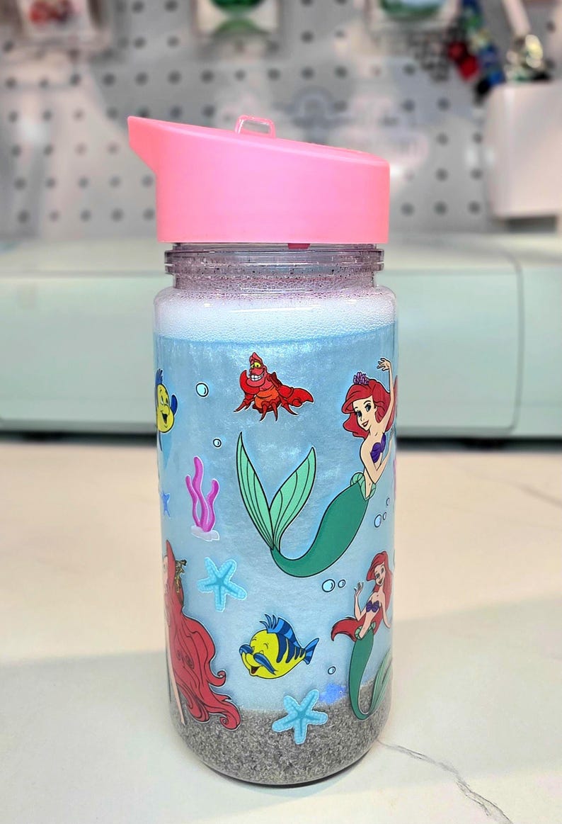 Może przedstawiać: Przezroczysta butelka na wodę z r&oacute;żową pokrywką z motywem syrenki. Butelka ma jasnoniebieskie tło z kresk&oacute;wkowymi ilustracjami syren, ryb i innych stworzeń morskich. Butelka jest wypełniona niebieskim płynem i piaszczystym dnem.