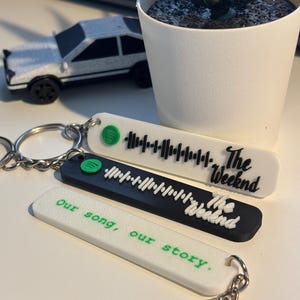May include: Three keychains with Spotify-style audio waveforms. Two keychains are white, one black. The white keychains have the text "The Weeknd" and "Our song, our story." The black keychain also has "The Weeknd" text. Each keychain has a silver keyring.