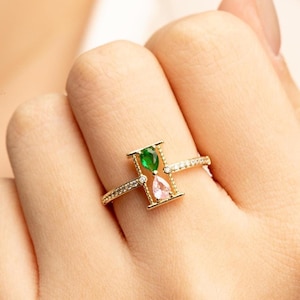 May include: A gold-colored ring featuring an hourglass design. The hourglass is set with a green and pink gemstone. The ring band is encrusted with small, clear gemstones, adding sparkle to the jewelry.