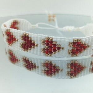 May include: White beaded bracelet featuring a pattern of red heart shapes, with gold-coloured beads. The bracelet is adjustable and is displayed on a reflective surface.
