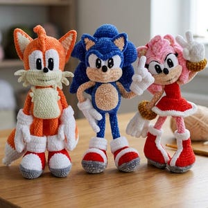 May include: Three handmade crochet plush dolls representing characters from the Sonic the Hedgehog series. The left doll is orange and cream, the center doll is blue and tan, and the right doll is pink, red, and white, all featuring yarn textures.