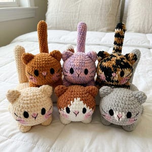 May include: Six crocheted cat plushies arranged in two rows. The cats feature round heads, upright tails, black oval eyes, pink stitched noses, and pink blush accents. The colors include cream, brown, lavender, tortoiseshell, and light gray yarn.