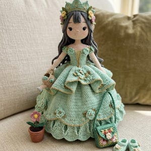 Könnte beinhalten: Eine handgefertigte Häkelpuppe mit dunkelgrauem Haar, die ein mintgrünes Kleid mit kleinen Blumen und eine passende Krone trägt. Die Puppe hält eine winzige Gießkanne und wird von einer kleinen Topfblume, einer grünen Handtasche und zwei Schuhen begleitet.