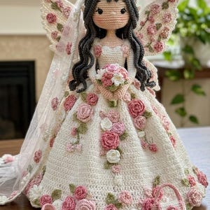 Könnte beinhalten: Eine gehäkelte Puppe mit langem dunklem Haar, die ein weißes Kleid mit rosa und weißen Blumenapplikationen, einen passenden Kopfschmuck und Flügel trägt. Die Puppe hält einen kleinen Blumenstrauß und wird von einem Paar Schuhe und einer Handtasche begleitet.