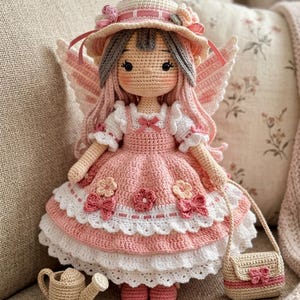 Könnte beinhalten: Eine handgefertigte Häkelpuppe mit langem rosa Haar, einem breitkrempigen Hut mit Blumenakzenten und Feenflügeln. Die Puppe trägt ein mehrlagiges rosa-weißes Kleid mit kleinen Blumenverzierungen sowie eine kleine Häkelgießkanne und eine Handtasche.