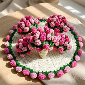 Könnte beinhalten: Ein kreisförmiges, weißes Häkeldeckchen mit einem grünen Rand, der mit kleinen rosa und hellrosa gehäkelten Blumen verziert ist. In der Mitte des Deckchens befinden sich vier Büschel aus gehäkelten rosa und hellrosa Rosen mit grünen Blättern.