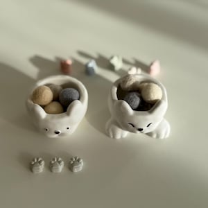 May include: Two white ceramic cat-shaped bowls filled with beige and gray felt balls. Three small paw-shaped ceramic pieces are in front of the bowls. Several small, colorful decorative pieces are in the background.