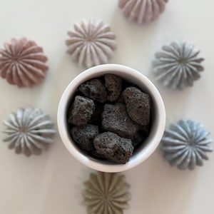 May include: A white bowl filled with dark gray lava rocks, surrounded by decorative, flower-shaped stones in shades of pink, gray, and green. The arrangement is set against a white background, creating a minimalist aesthetic.