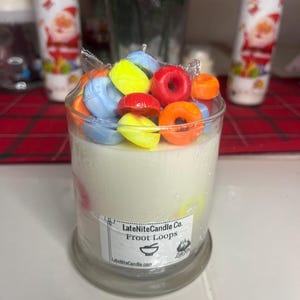 Op de afbeelding: Een cilindrische kaars met een witte wasbasis, bekroond met kleurrijke, ringvormige stukjes was. De kaars zit in een doorzichtige glazen container. Het label luidt "LateNiteCandle Co. Froot Loops".