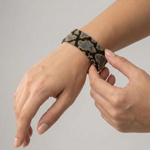 May include: A beaded bracelet with a snake skin pattern in black, gray, and gold. The bracelet is wide and wraps around the wrist. The clasp is gold. The bracelet is being clasped on a person's wrist.