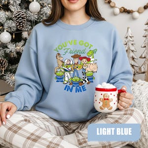May include: Light blue sweatshirt featuring a graphic of characters from the movie Toy Story. The text "You've Got a Friend in Me" is printed above the characters. A white mug with a gingerbread man design is held in the hand.