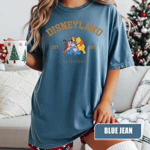 Pu&ograve; includere: T-shirt blu jeans con la scritta "DISNEYLAND" in lettere dorate. La maglietta presenta una grafica di Winnie the Pooh, Tigro, Pimpi ed Eeyore. Il testo "EST. 1955 CALIFORNIA" &egrave; stampato anche sulla maglietta.