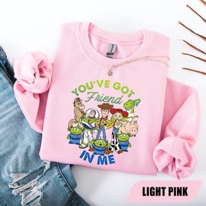 May include: Light pink sweatshirt featuring a graphic of characters from the movie Toy Story. The text "YOU'VE GOT A FRIEND IN ME" is above the characters. The sweatshirt is made of a soft material and is a crew neck style.