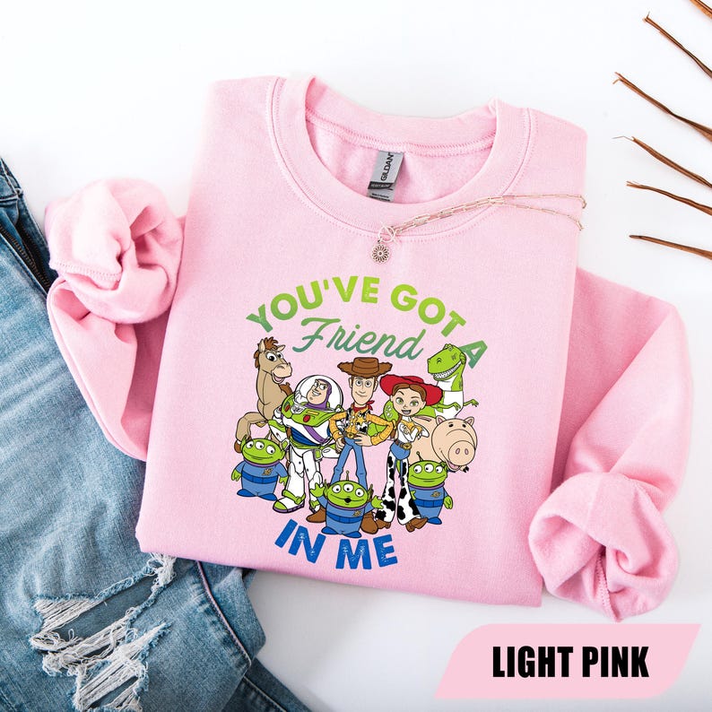May include: Light pink sweatshirt featuring a graphic of characters from the movie Toy Story. The text "YOU'VE GOT A FRIEND IN ME" is above the characters. The sweatshirt is made of a soft material and is a crew neck style.