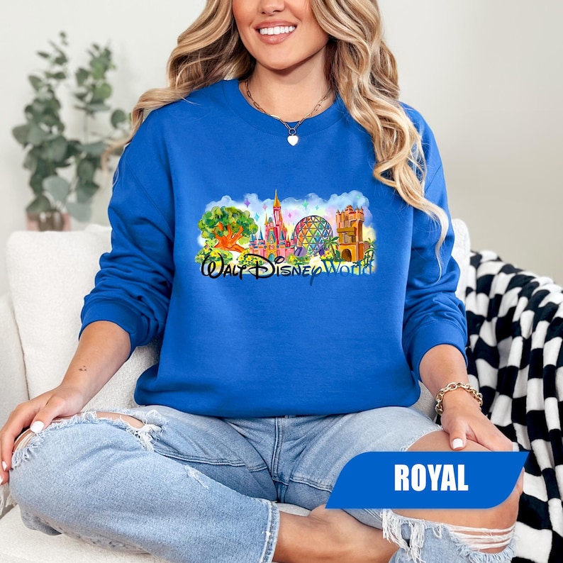 May include: Royal blue sweatshirt featuring a colorful graphic of the Walt Disney World logo. The graphic includes a tree, castle, Spaceship Earth, and other park landmarks. The sweatshirt is long-sleeved and made of a soft material.