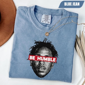 May include: Blue jean Comfort Colors t-shirt featuring a graphic of a person's face with the text "BE HUMBLE" in a red rectangle. A gold necklace with a pendant is draped on the shirt.