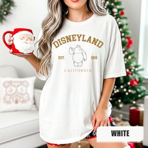 Pu&ograve; includere: T-shirt bianca con grafica dorata di Disneyland. Il design include il testo "Disneyland", "Est. 1955" e "California", insieme a un'illustrazione di un personaggio. La maglietta &egrave; di vestibilit&agrave; ampia.