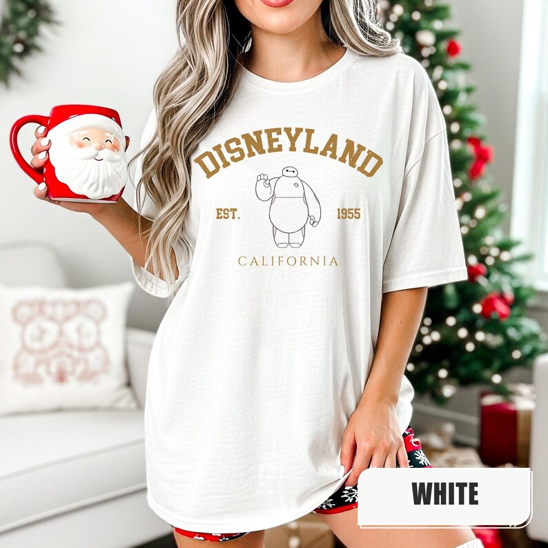 Pu&ograve; includere: T-shirt bianca con grafica dorata di Disneyland. Il design include il testo "Disneyland", "Est. 1955" e "California", insieme a un'illustrazione di un personaggio. La maglietta &egrave; di vestibilit&agrave; ampia.