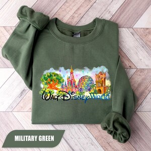 May include: Military green sweatshirt featuring a colorful graphic of the Walt Disney World castle, Spaceship Earth, and other park landmarks. The text "Walt Disney World" is below the graphic. The sweatshirt is folded on a light wood surface.