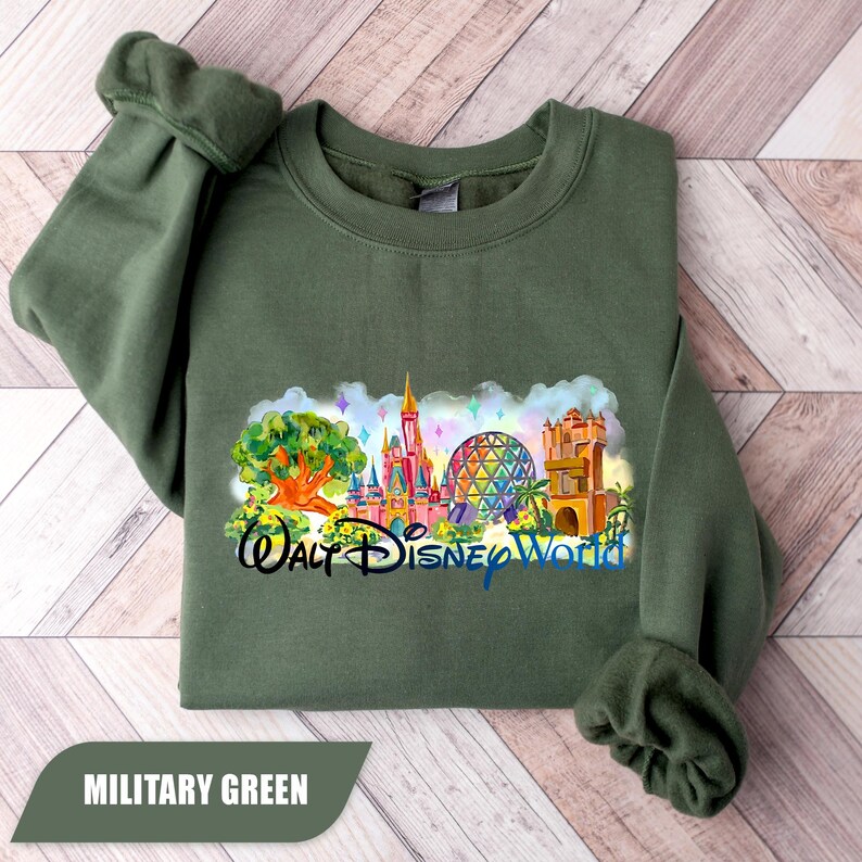 May include: Military green sweatshirt featuring a colorful graphic of the Walt Disney World castle, Spaceship Earth, and other park landmarks. The text "Walt Disney World" is below the graphic. The sweatshirt is folded on a light wood surface.