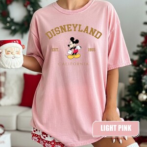 Pu&ograve; includere: T-shirt rosa chiaro con la scritta "Disneyland" in lettere dorate, insieme a "Est. 1955" e "California". Una grafica di Topolino &egrave; centrata sotto la parola "Disneyland". La maglietta &egrave; di colore rosa chiaro.