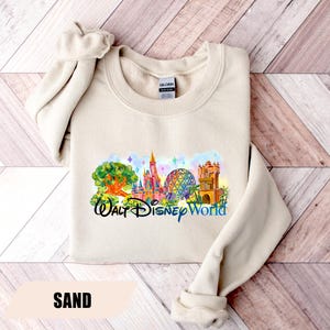 May include: Sand-colored sweatshirt featuring a colorful graphic of Walt Disney World landmarks. The design includes a castle, Spaceship Earth, and other attractions, with the words "Walt Disney World" in a script font. The sweatshirt has a crew neck and long sleeves.