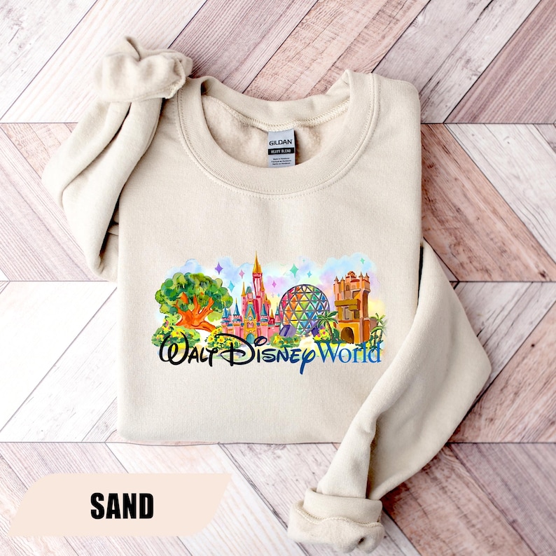 May include: Sand-colored sweatshirt featuring a colorful graphic of Walt Disney World landmarks. The design includes a castle, Spaceship Earth, and other attractions, with the words "Walt Disney World" in a script font. The sweatshirt has a crew neck and long sleeves.